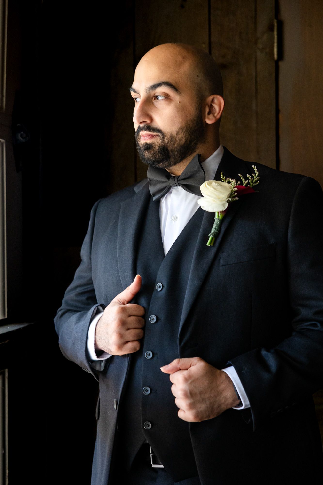 Bald groom with beard in dark suit, bow tie, and white rose boutonniere gazes thoughtfully toward window light.