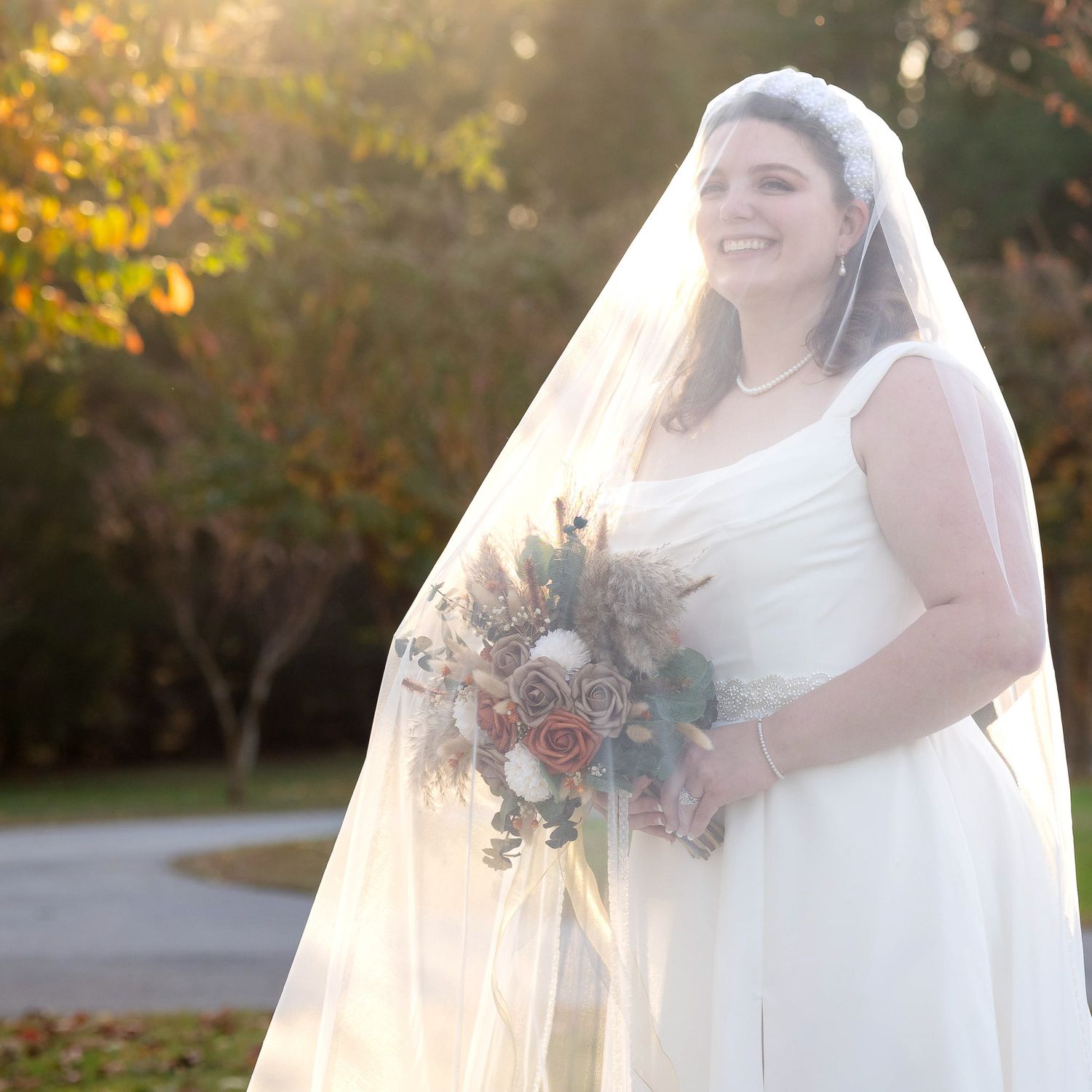 Smiling bride in white gown and long veil holds autumn bouquet outdoors in warm golden sunlight.