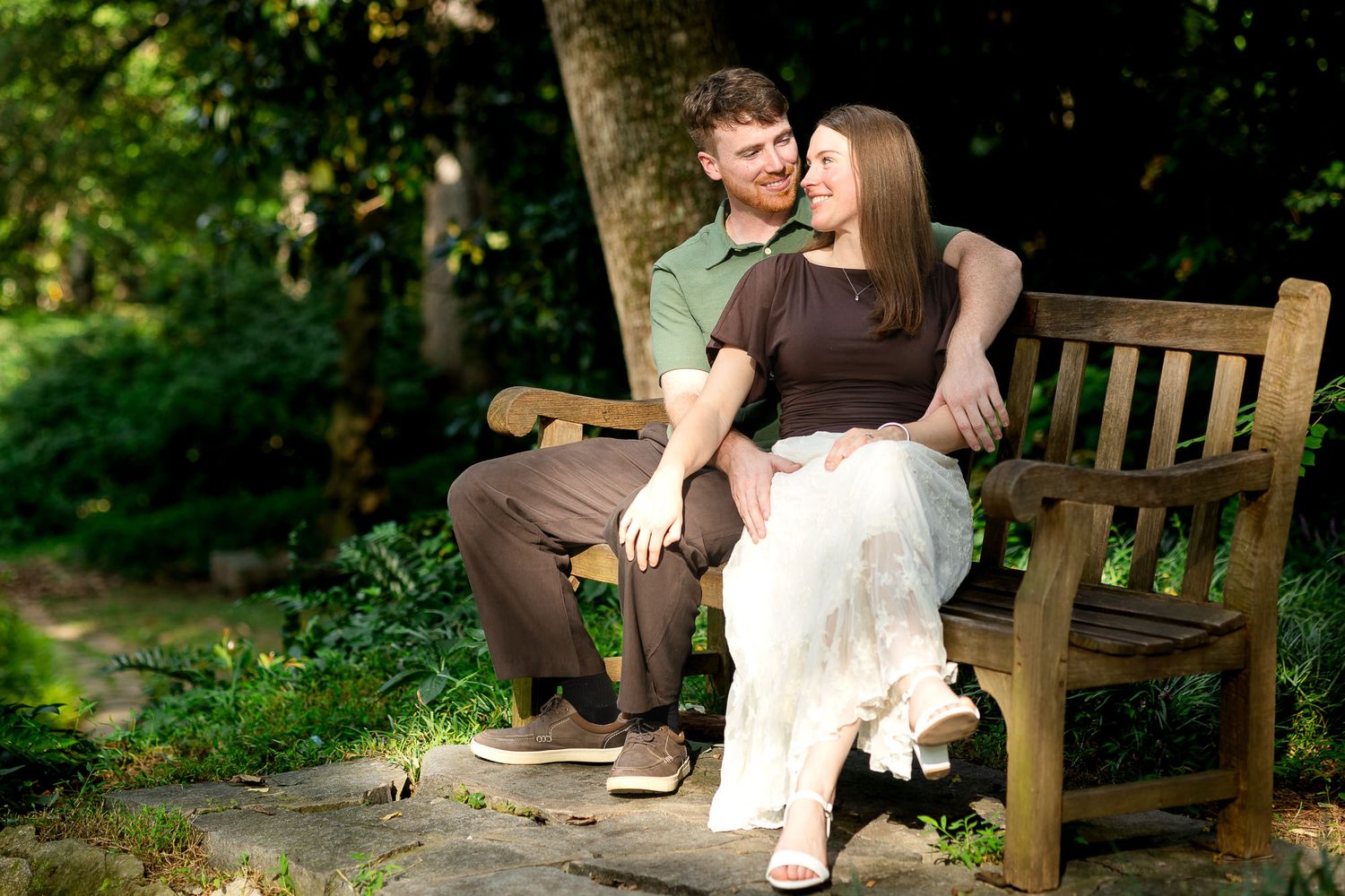 Couple sitting closely on a wooden bench in a lush green garden, smiling and enjoying a romantic outdoor moment.