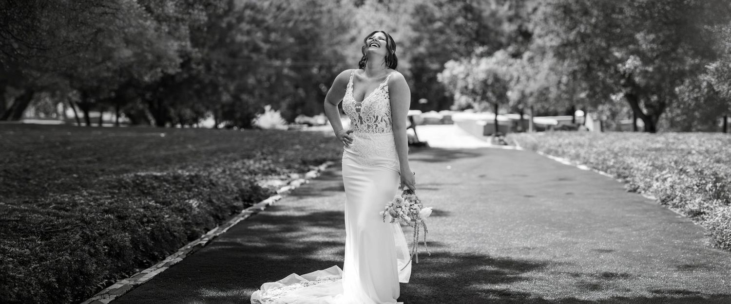 Black and white photo of a bride in a fitted wedding gown walking down a tree-lined path holding a bouquet.