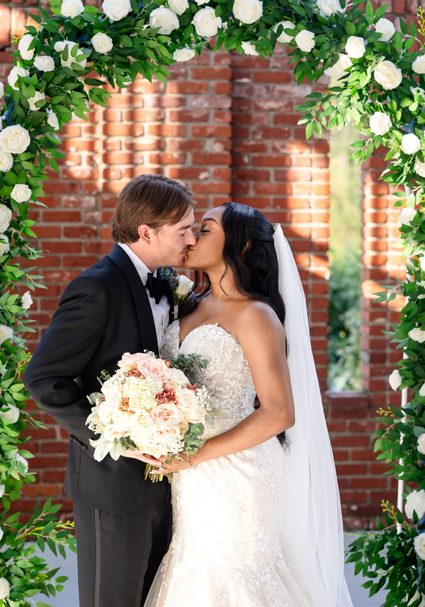 Bride and groom share a kiss under a floral arch adorned with white roses and greenery at their outdoor wedding ceremony.