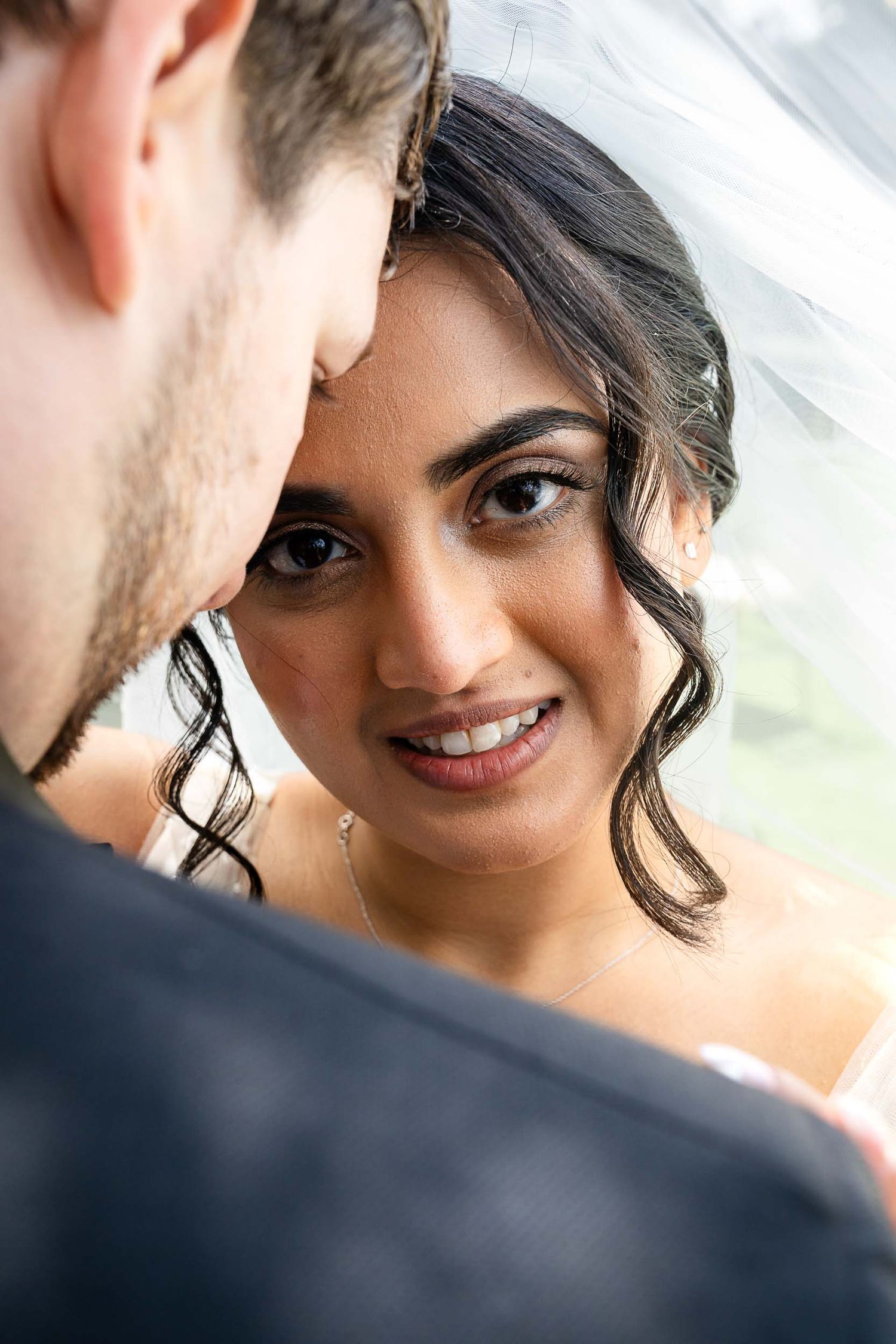 Radiant bride smiles at groom under wedding veil during romantic portrait session.