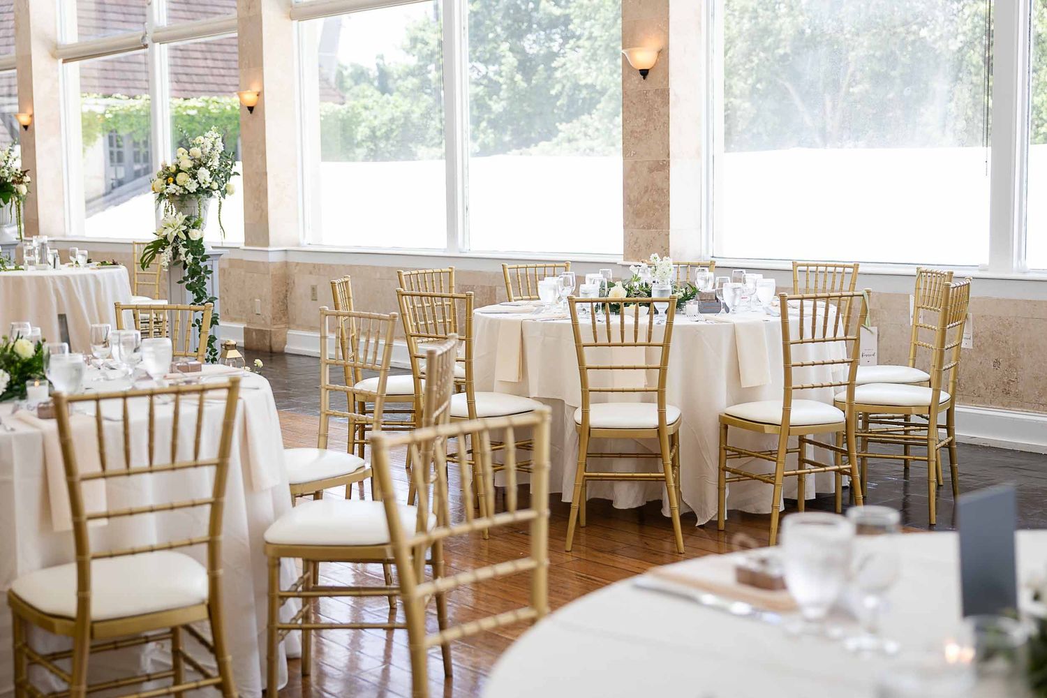 Elegant wedding reception hall with gold Chiavari chairs, white round tables, floral centerpieces, and large windows with natural light.