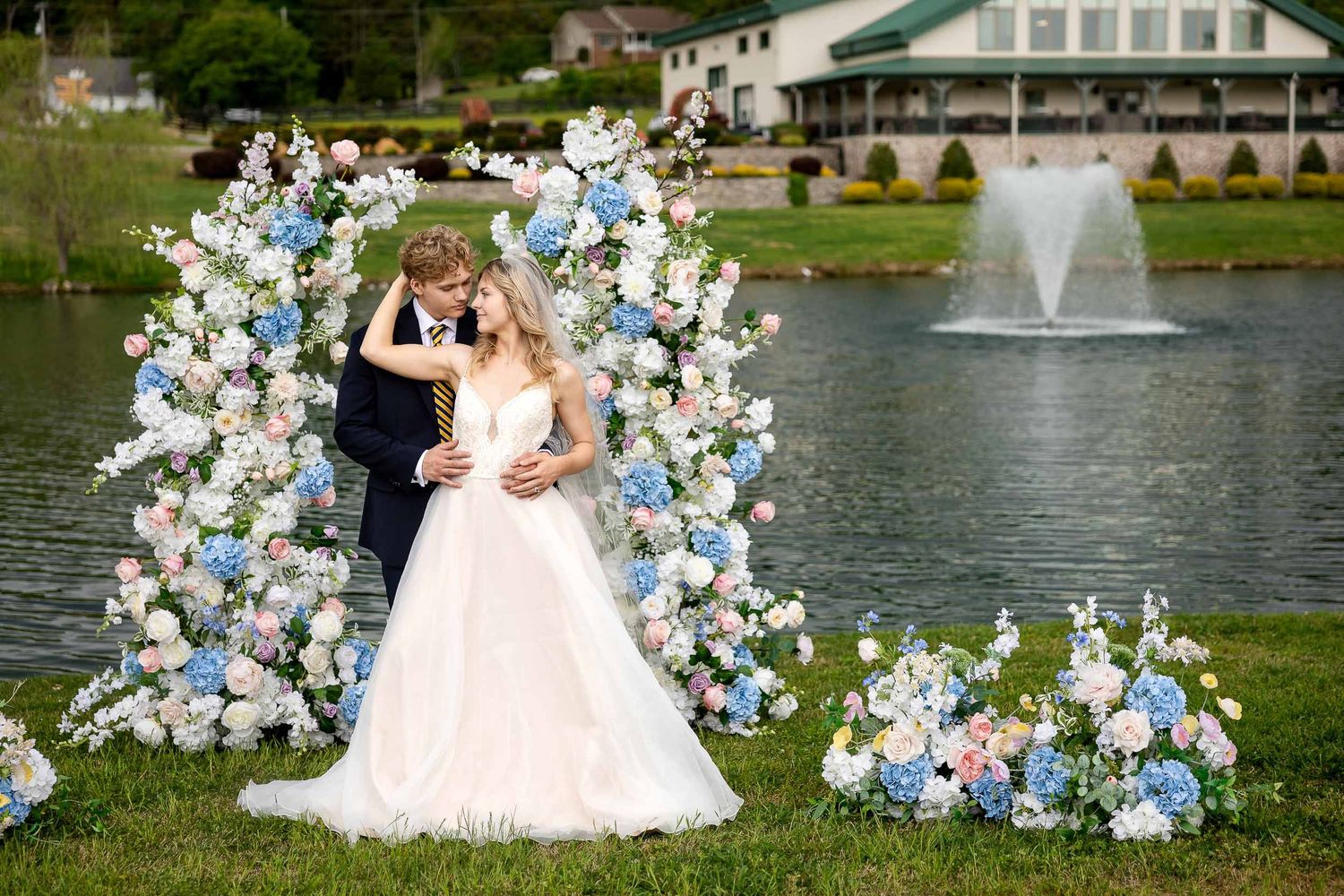 A wedding couple stands beneath a floral arch of blue and white blooms beside a lake with a fountain in the background.