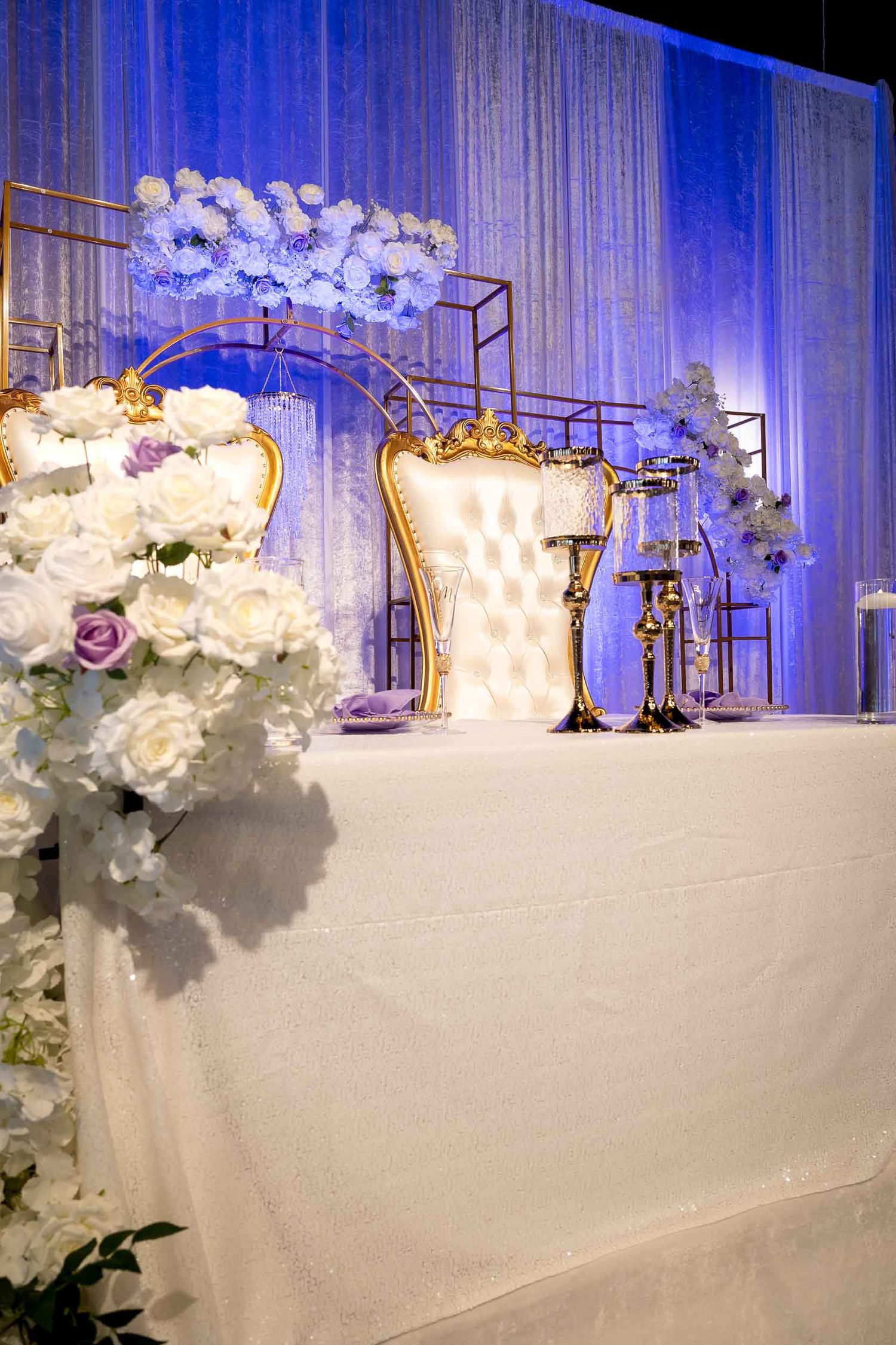 Elegant wedding sweetheart table with white floral arrangements, gold throne chairs, and purple uplighting backdrop.