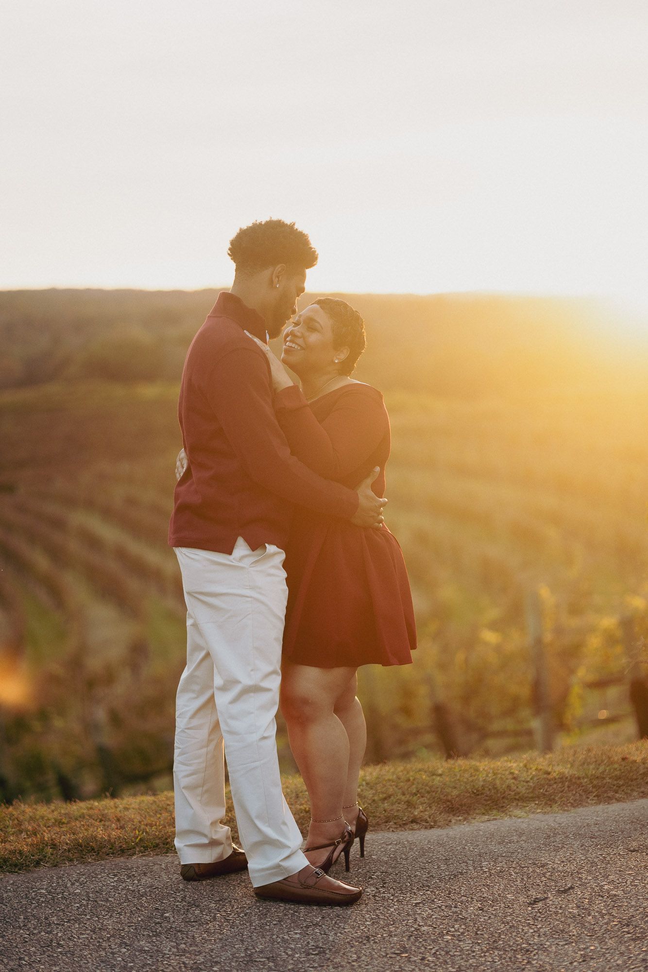 Couple embracing and smiling at each other on a quiet road at golden hour, with sunlit vineyard hills glowing behind them.