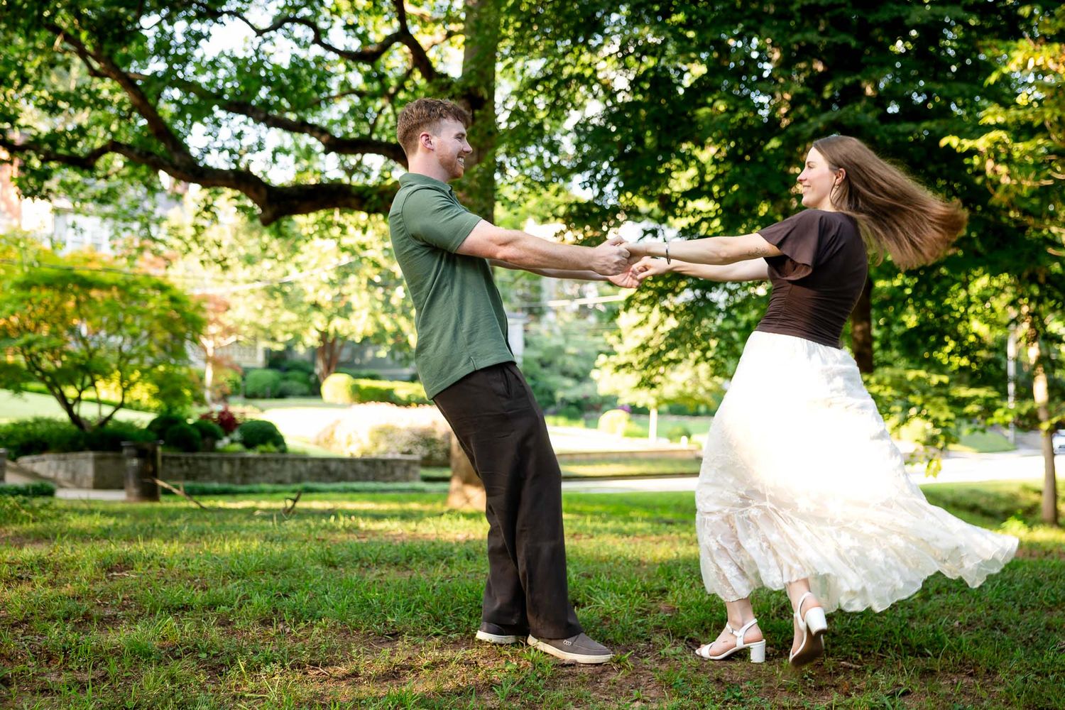 Couple dancing joyfully in a lush green park, woman in white skirt spinning with hair flowing.