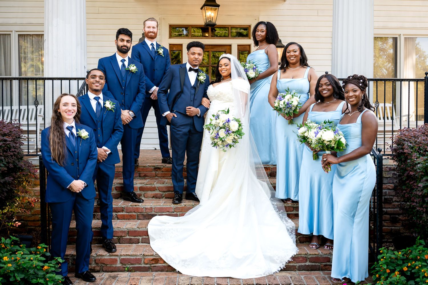 Wedding party poses on steps, bride in white gown with groomsmen in navy suits and bridesmaids in light blue dresses.