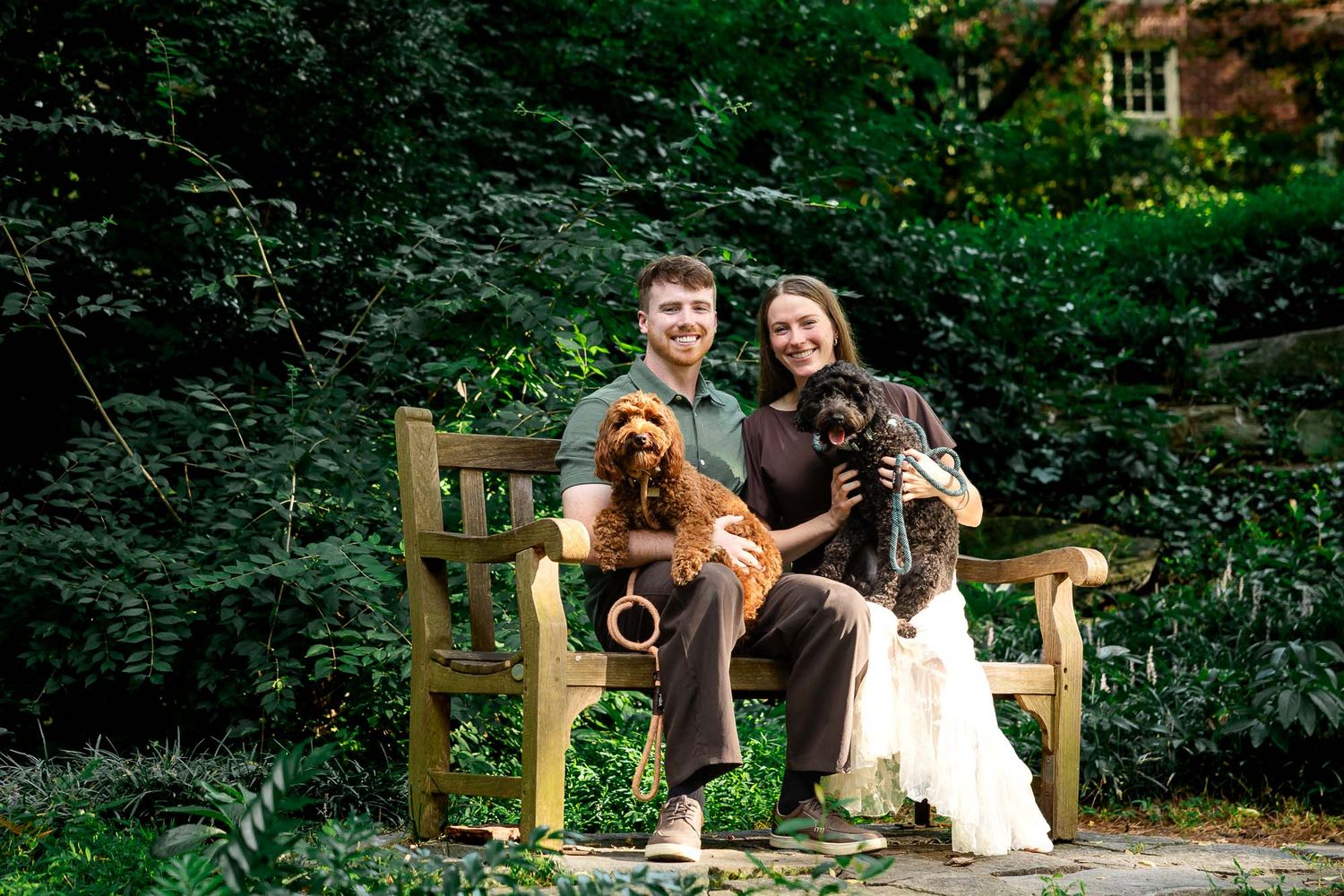 Smiling couple sitting on wooden garden bench holding two dogs surrounded by lush green foliage.