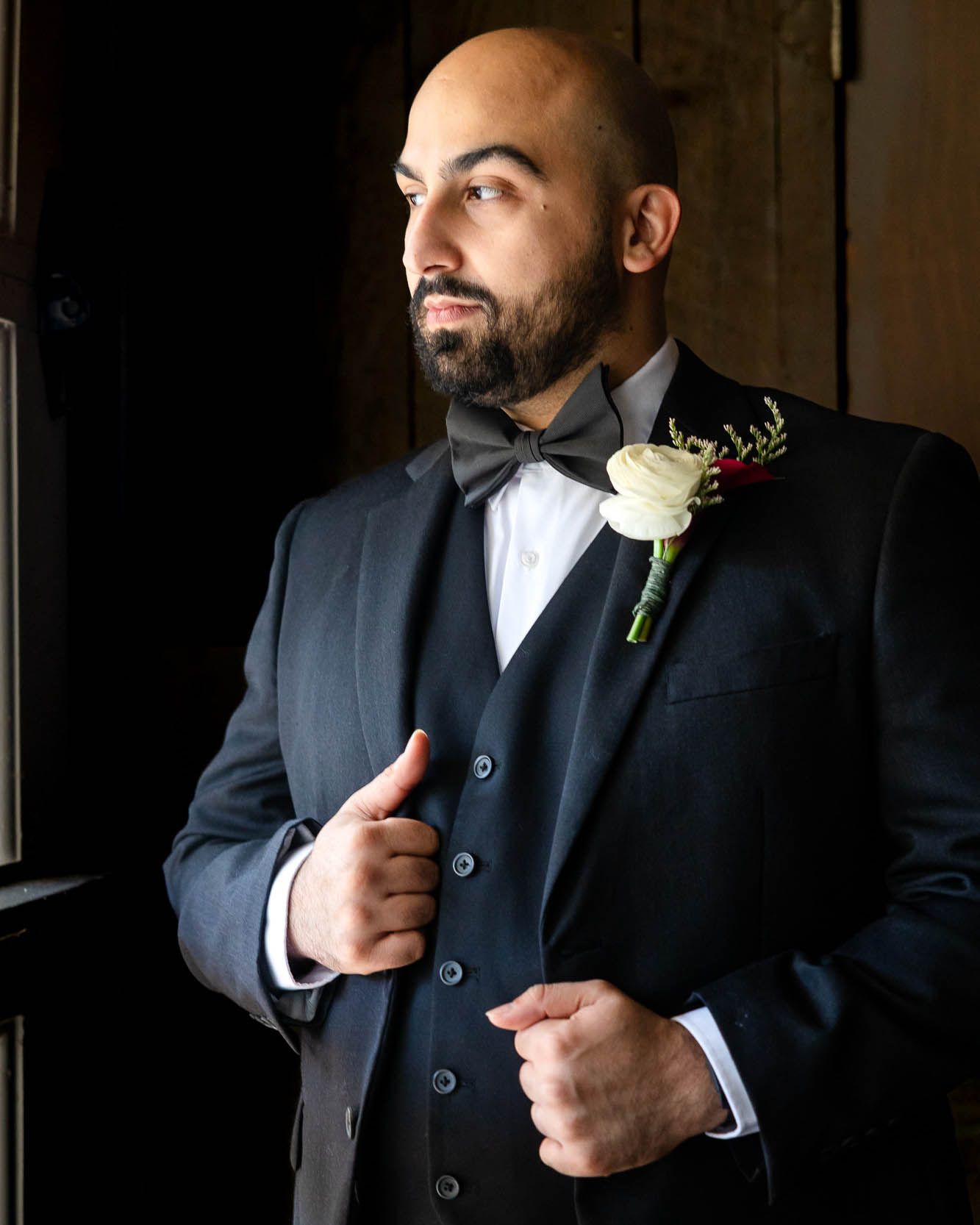 Bald groom in dark three-piece suit with bow tie and white rose boutonniere posing indoors.