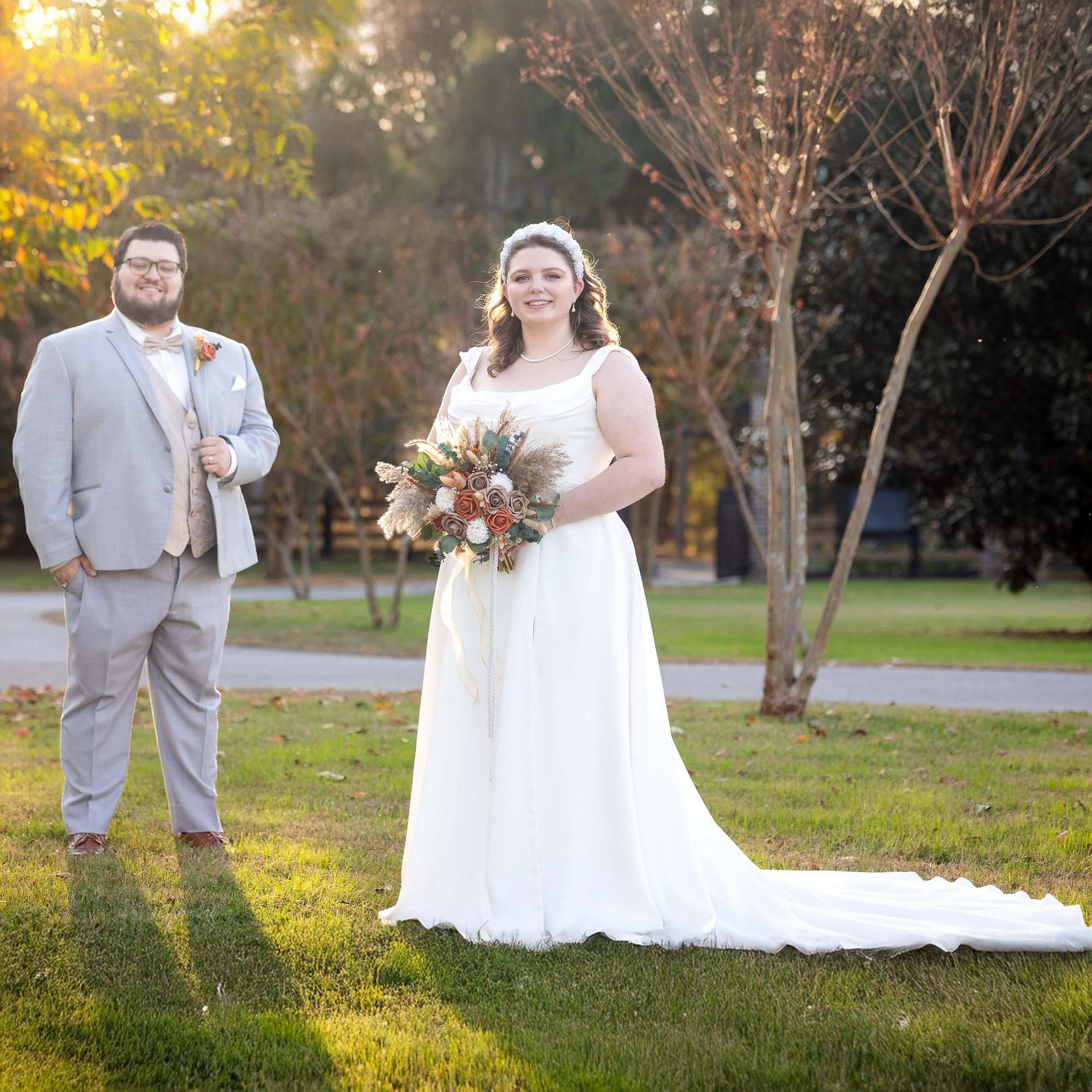 Bride in white gown holding bouquet stands with groom in gray suit on green lawn with autumn trees.