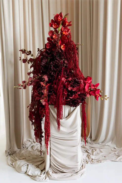 Dramatic deep red floral arrangement with orchids and amaranthus draped over an elegant ivory fabric pedestal.