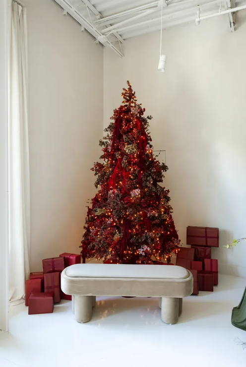 Red Christmas tree with fairy lights and wrapped gifts beside a white bench in a minimalist white room.
