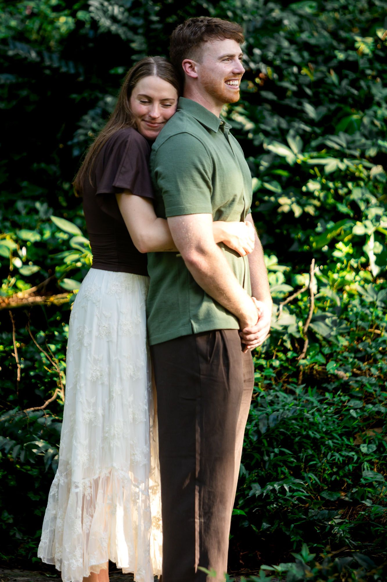 A couple embraces outdoors, woman in white skirt hugging man in green polo, surrounded by lush green foliage.