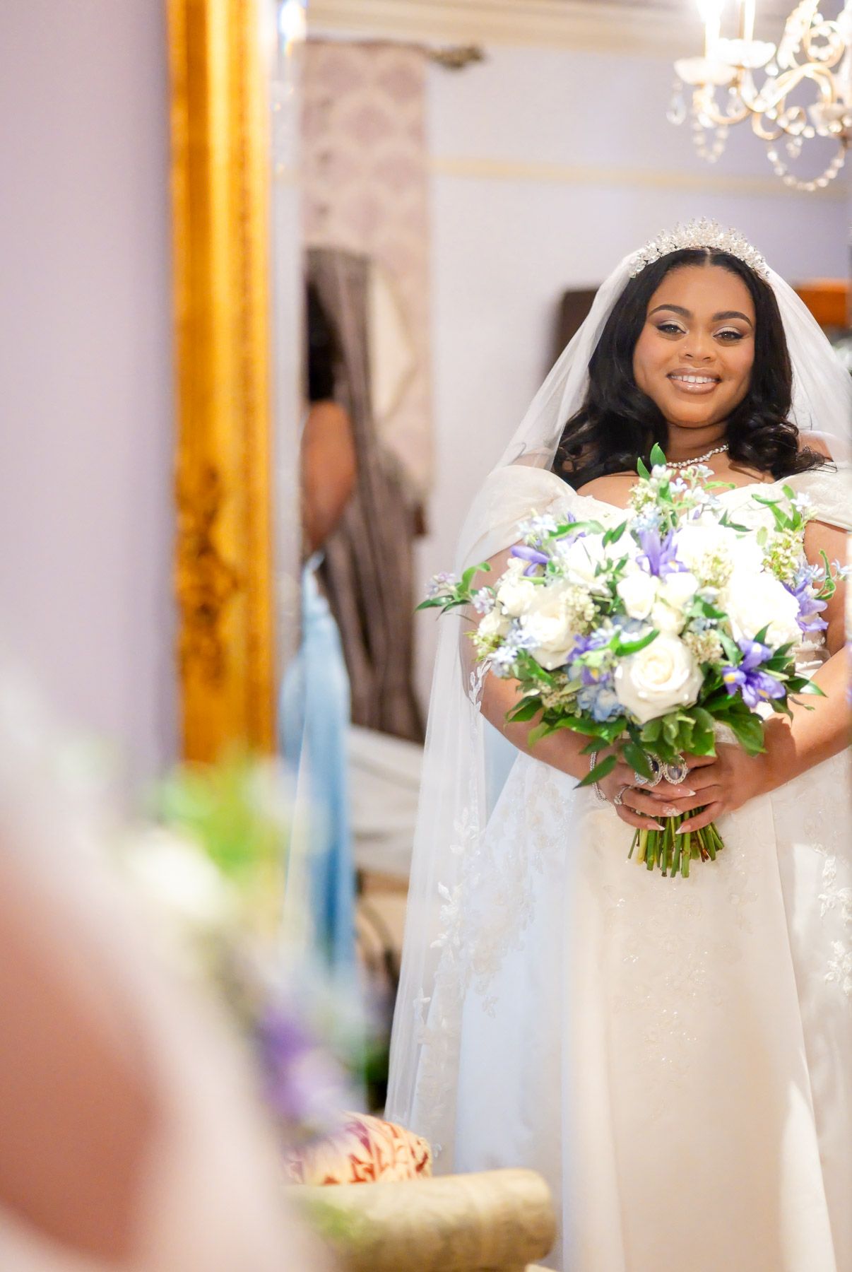 Smiling bride in white gown and veil holds purple and white bouquet, standing in elegant room with gold mirror.
