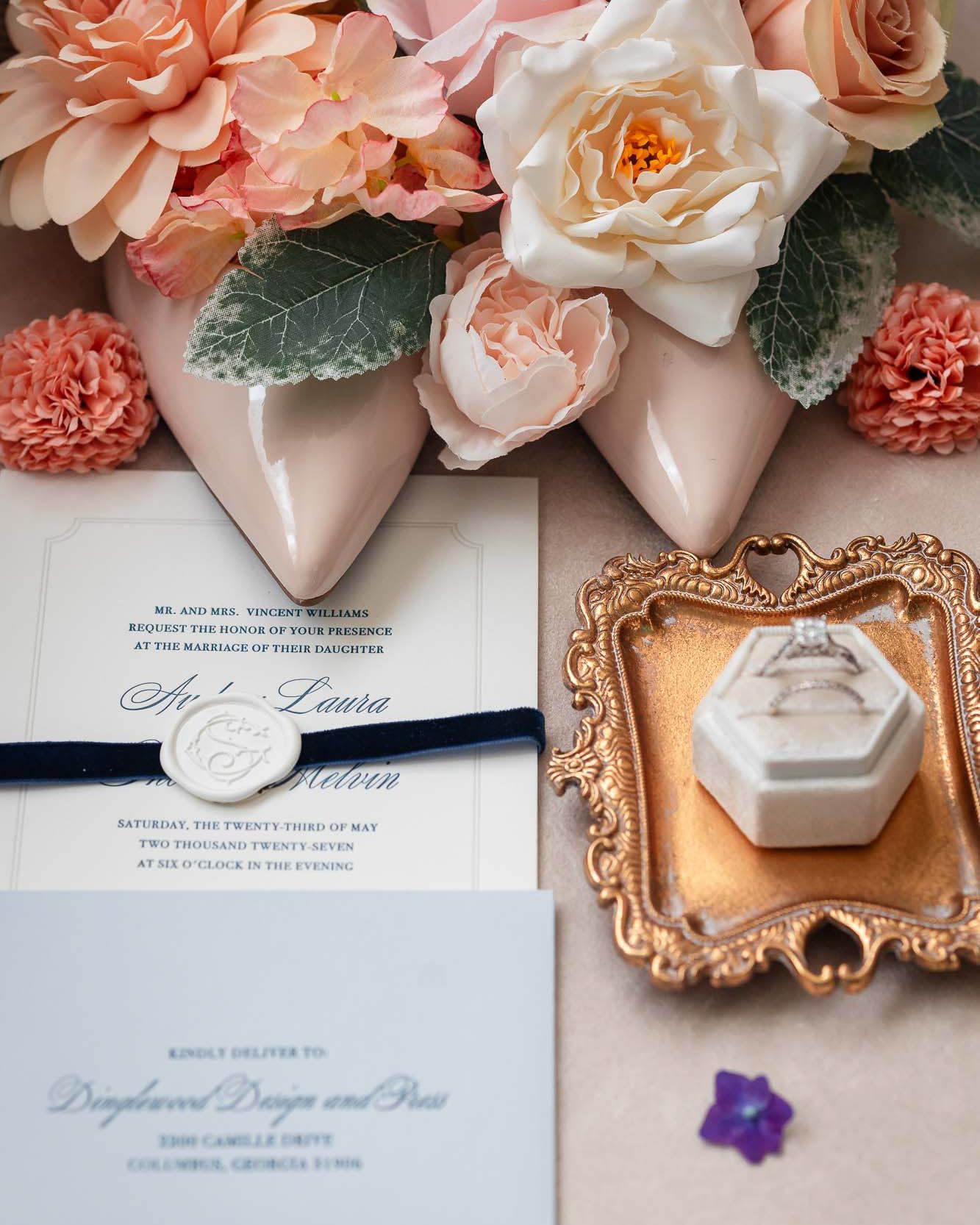 Elegant wedding flat lay featuring floral bouquet, blush heels, navy ribbon invitation, diamond ring in hexagon box on gold tray.