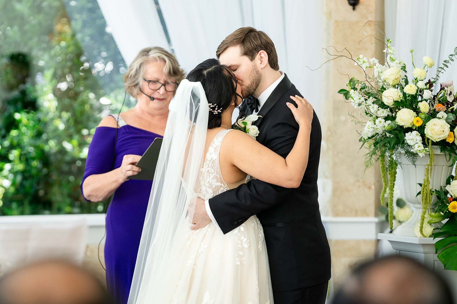 Bride and groom share first kiss at wedding ceremony altar with floral arrangements and guest in purple dress nearby.