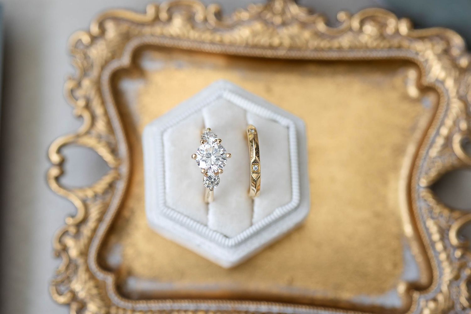 Two gold rings in a white hexagon velvet box on a gold ornate tray, featuring a diamond cluster engagement ring.
