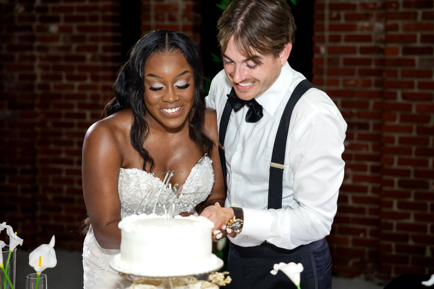 Newlywed couple cutting white wedding cake, bride in lace gown and groom in suspenders, smiling joyfully.