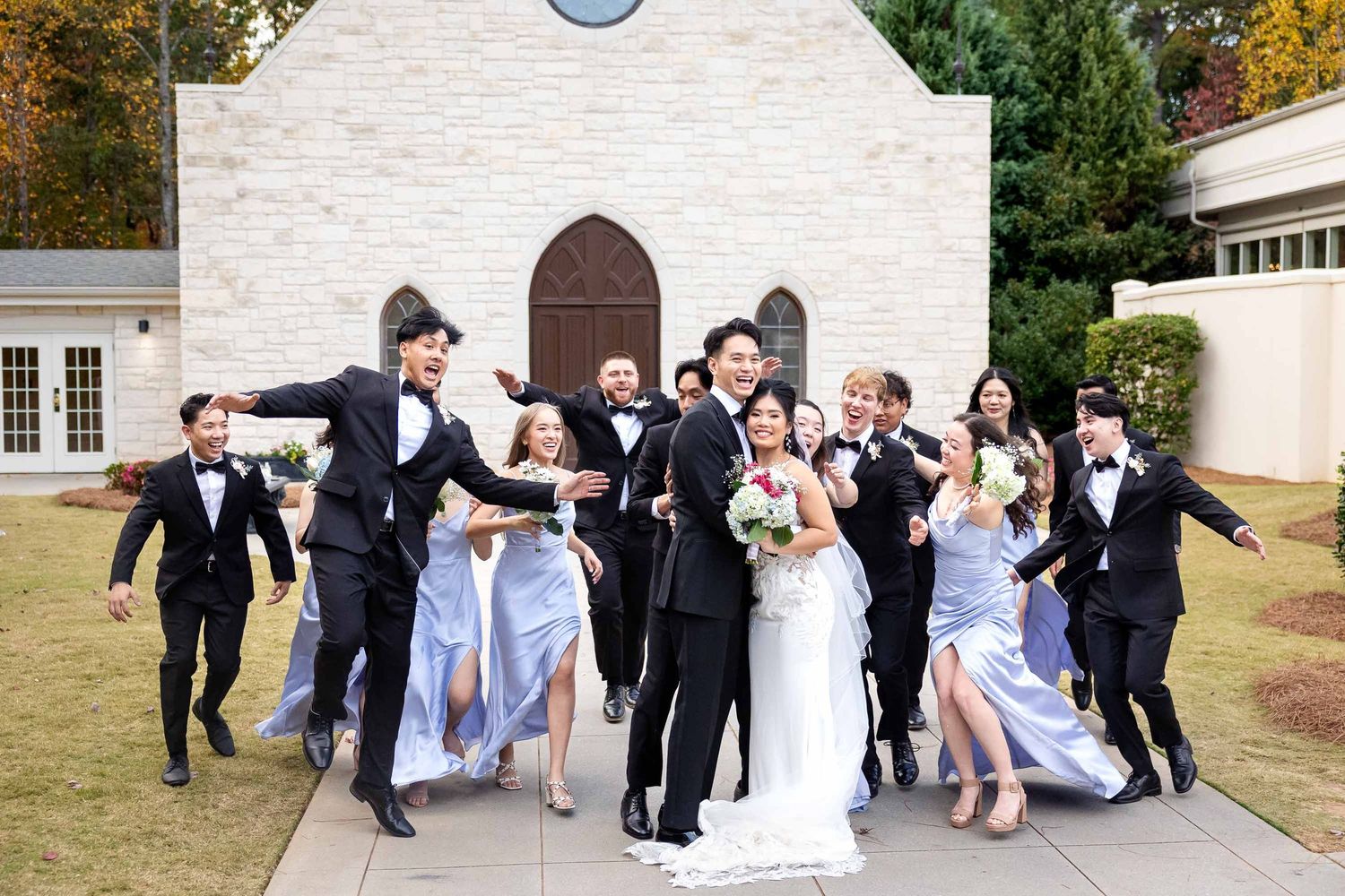 Wedding party celebrating outside a stone church, bridesmaids in blue dresses and groomsmen in black tuxedos.