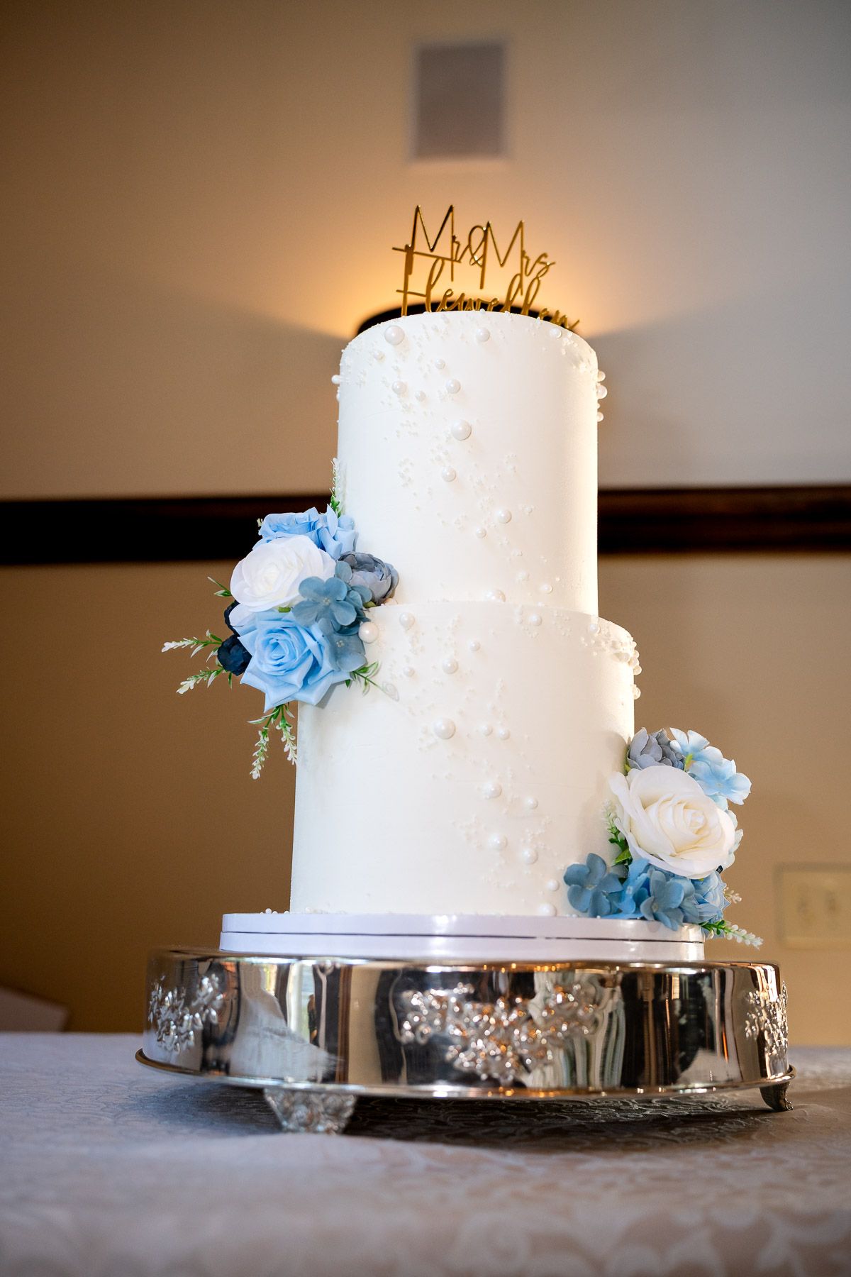 Elegant three-tier white wedding cake with blue and white floral accents and gold Mr & Mrs topper on silver stand.