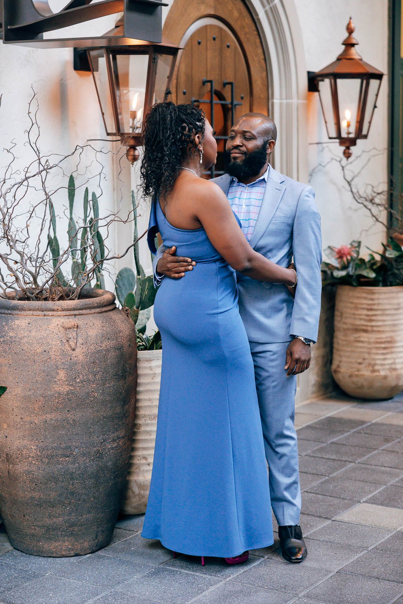 Couple in coordinated blue outfits embracing in a courtyard with vintage lanterns, large terracotta planters and an arched wooden door.
