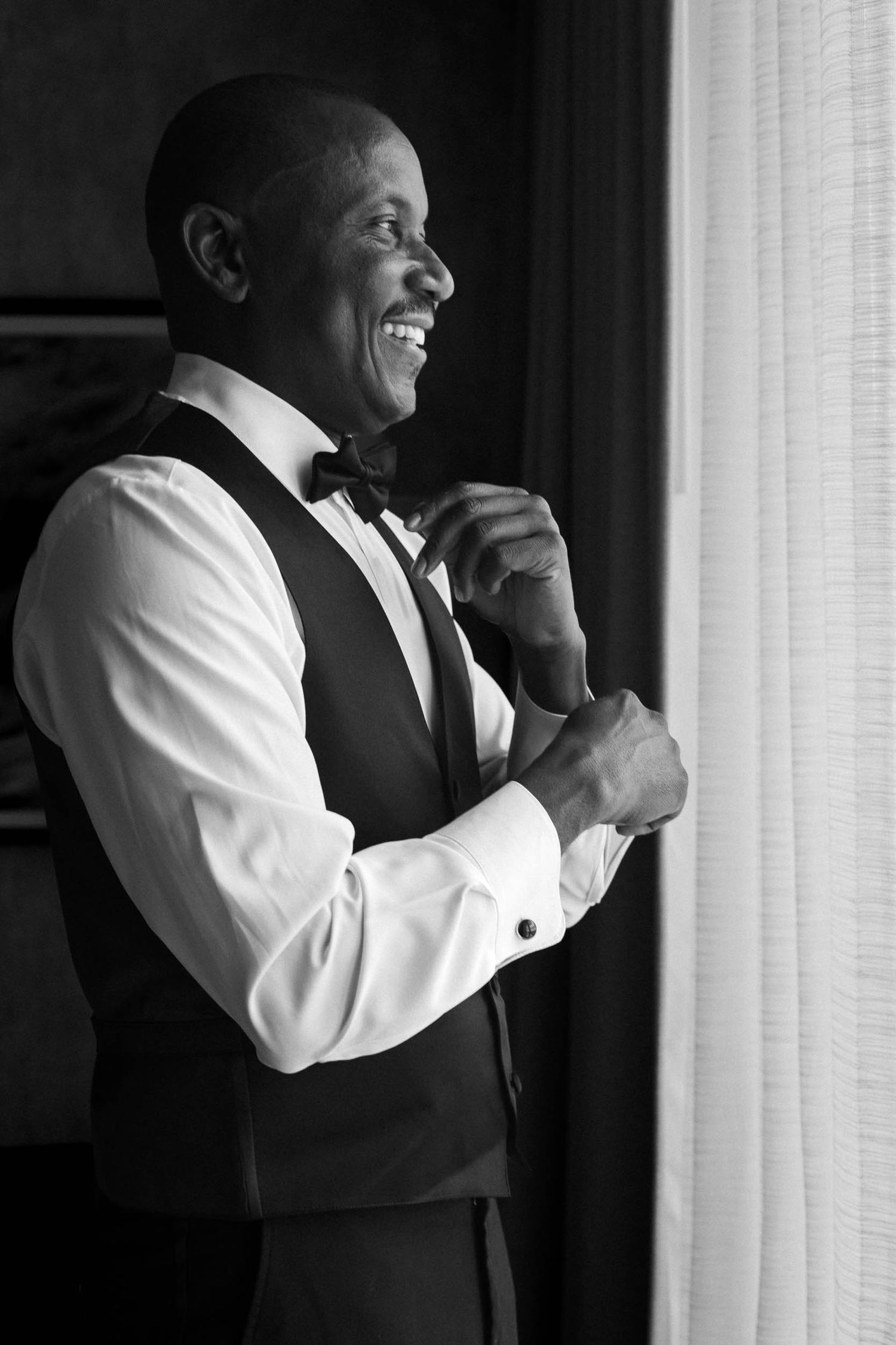 Smiling groom in black vest and bow tie adjusting cufflinks by a window in black and white photo.