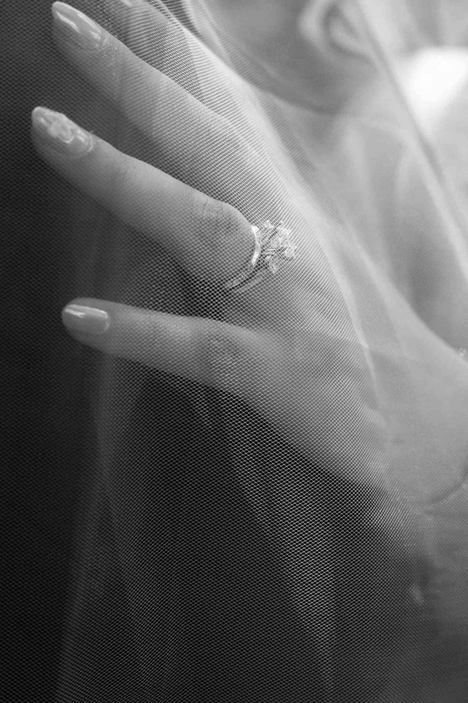 Black and white close-up of a bride's hand wearing a diamond engagement ring draped over a sheer wedding veil.