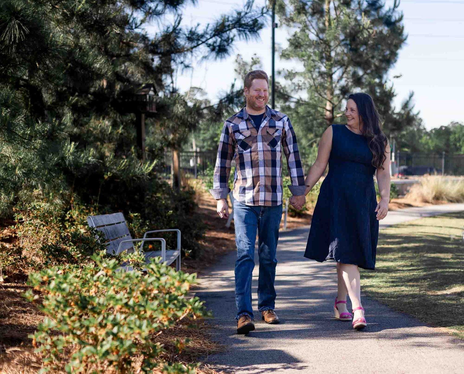 Couple holding hands walking on a sunny park path surrounded by pine trees and green landscaping.