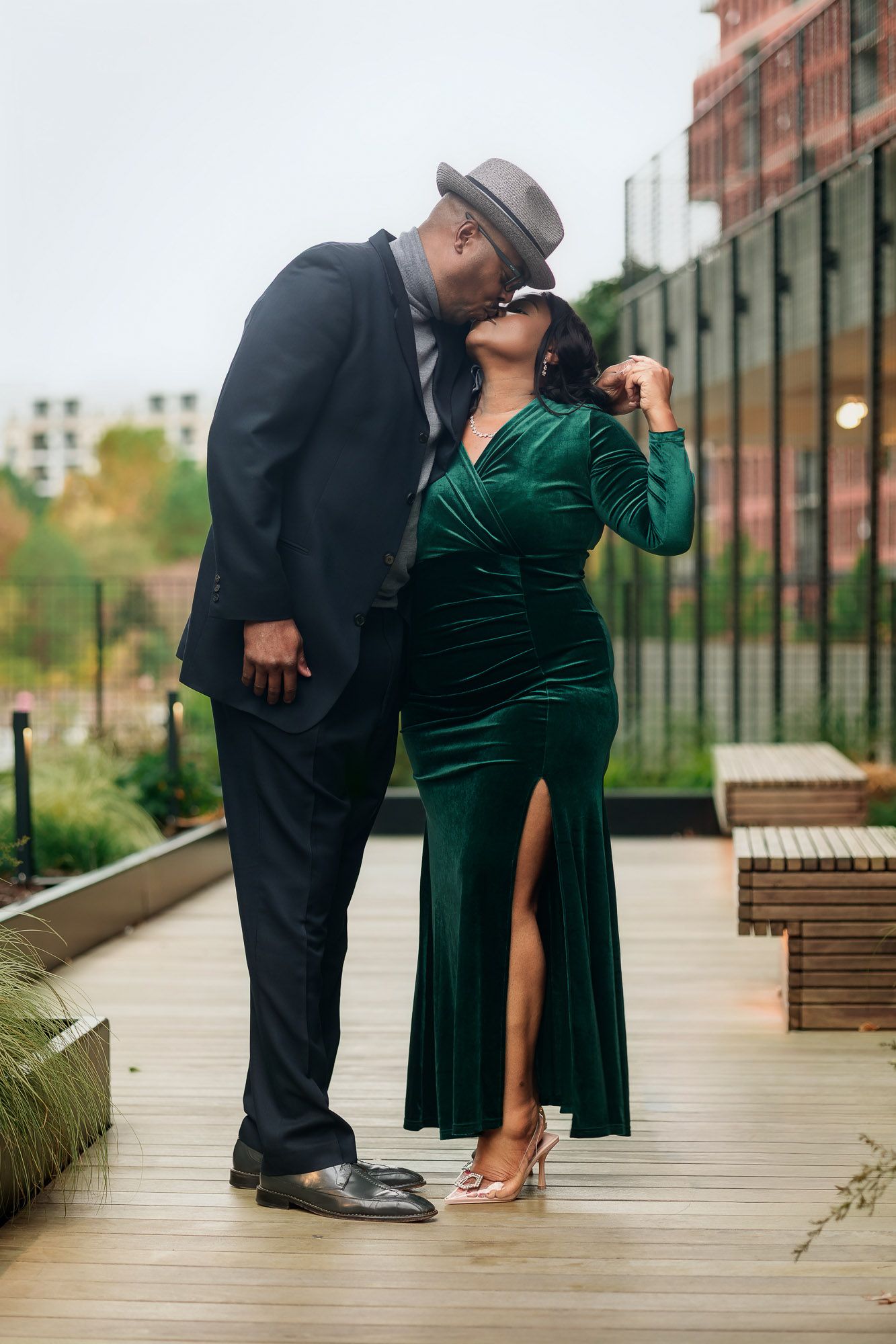Couple sharing a kiss outdoors, man in suit and hat, woman in elegant green velvet dress on a wooden deck.