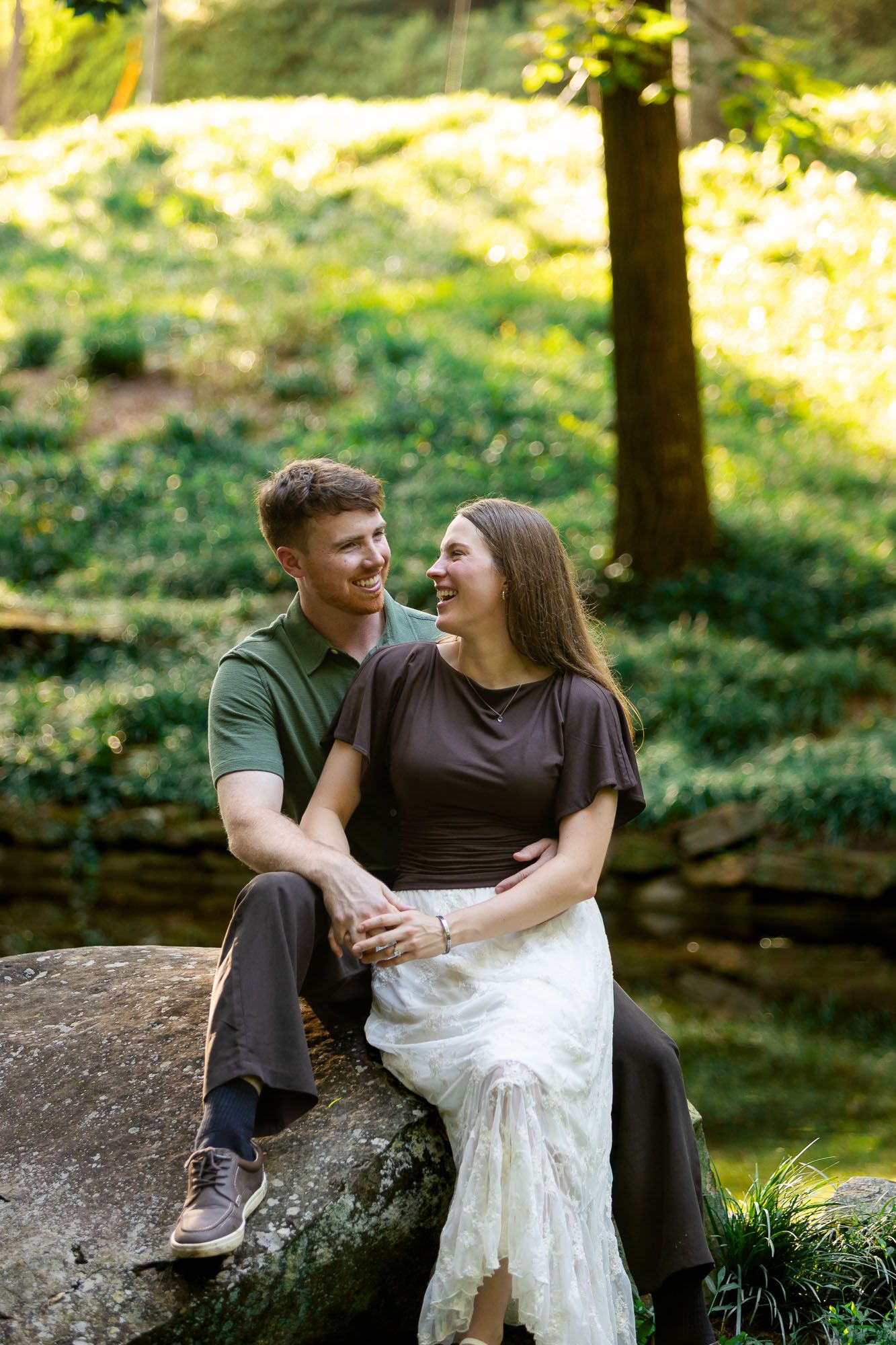 Couple sitting on a rock in a lush green garden, smiling at each other during an outdoor engagement photo session.