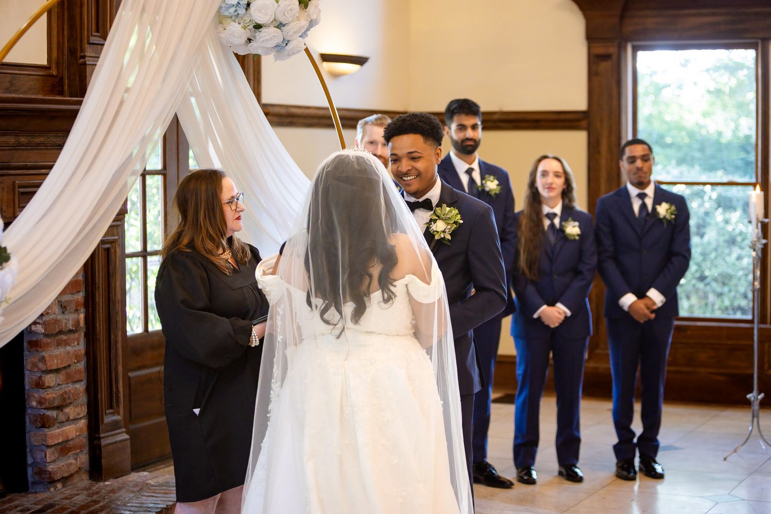 Bride and groom exchange vows at elegant indoor wedding ceremony with wedding party standing behind them.