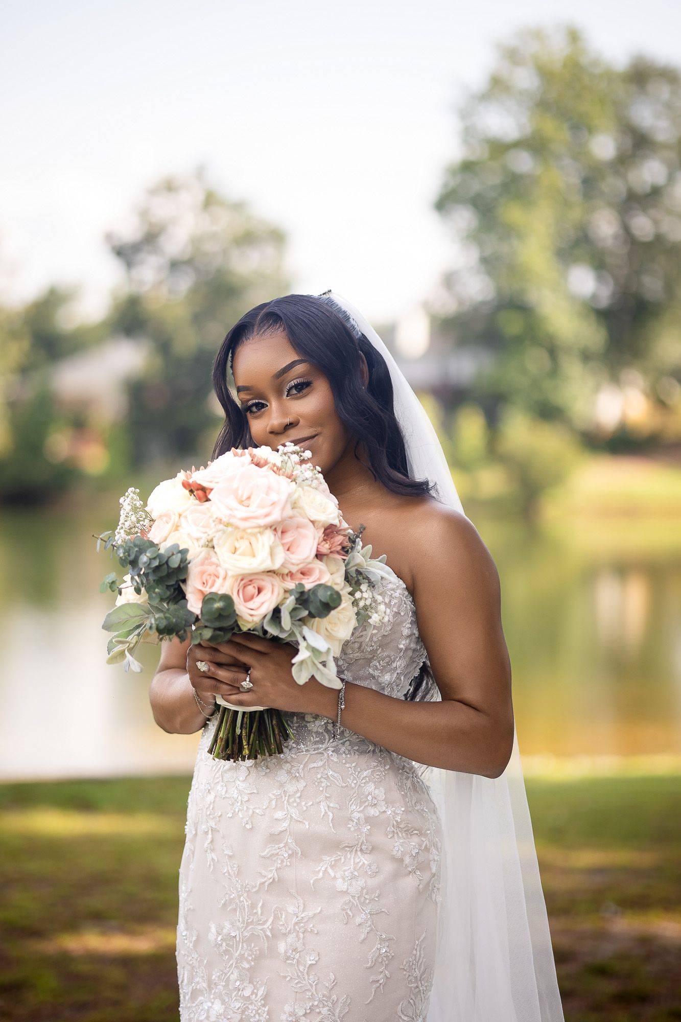 Beautiful Black bride in lace dress holding blush floral bouquet near serene lake.