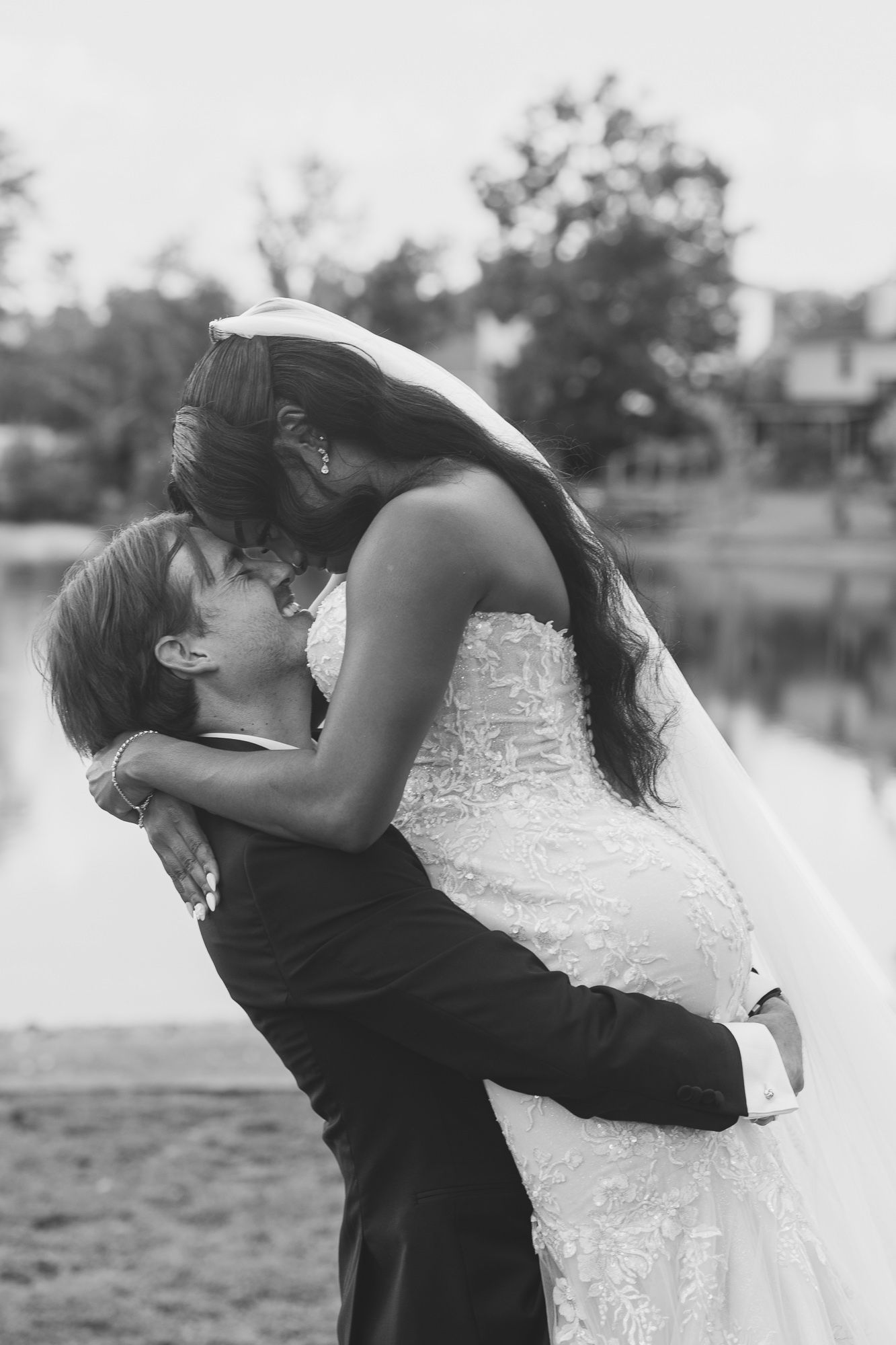 Black and white photo of a bride and groom sharing a romantic kiss outdoors near a lake on their wedding day.