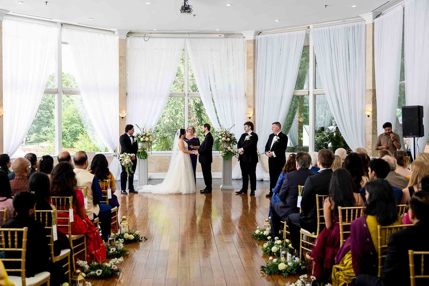 Elegant indoor wedding ceremony with white drapes, floral arrangements, and guests seated in gold chairs on hardwood floors.