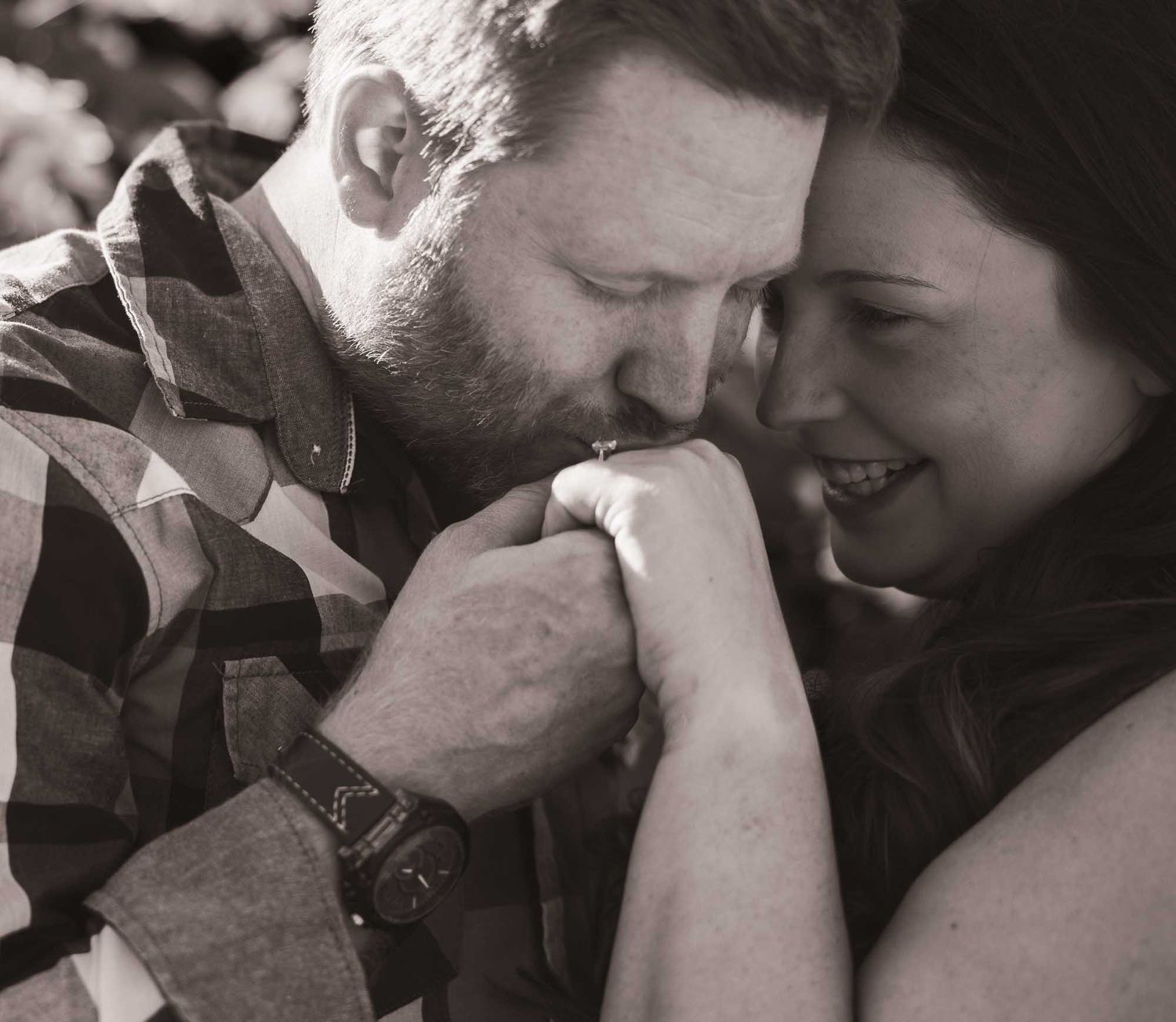 Black and white photo of a bearded man kissing a woman's hand as she smiles joyfully in a romantic moment.