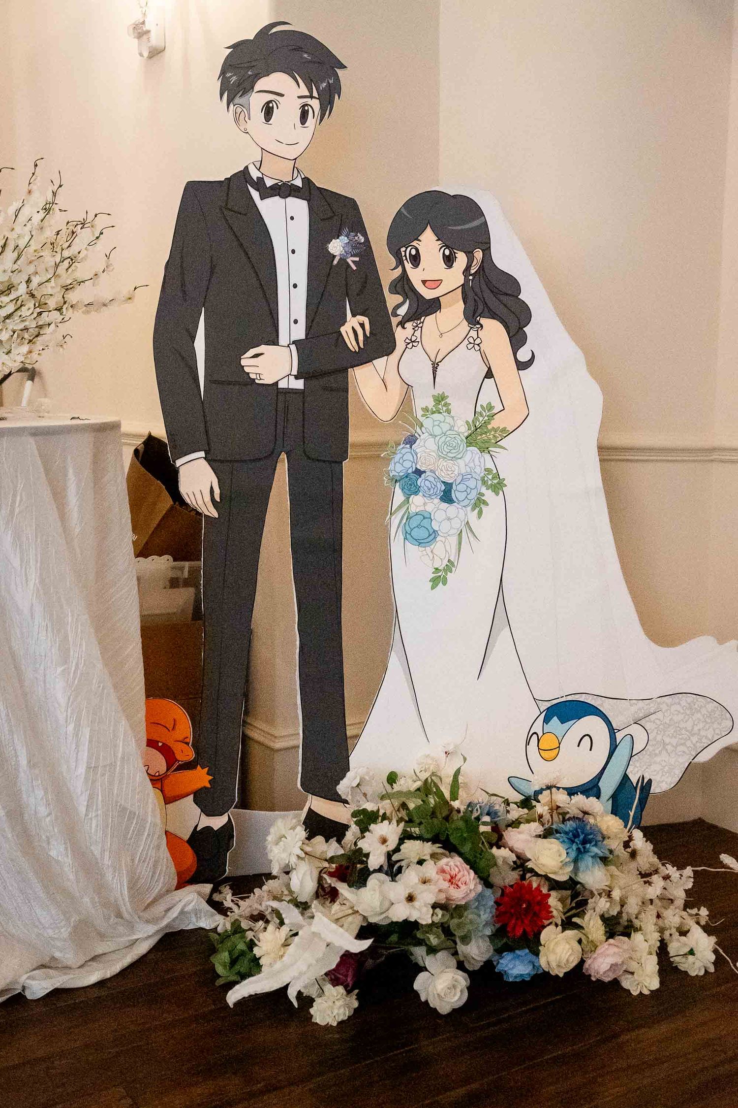 Anime-style cardboard cutout of a wedding couple in formal attire with Piplup and floral arrangements at a wedding venue.