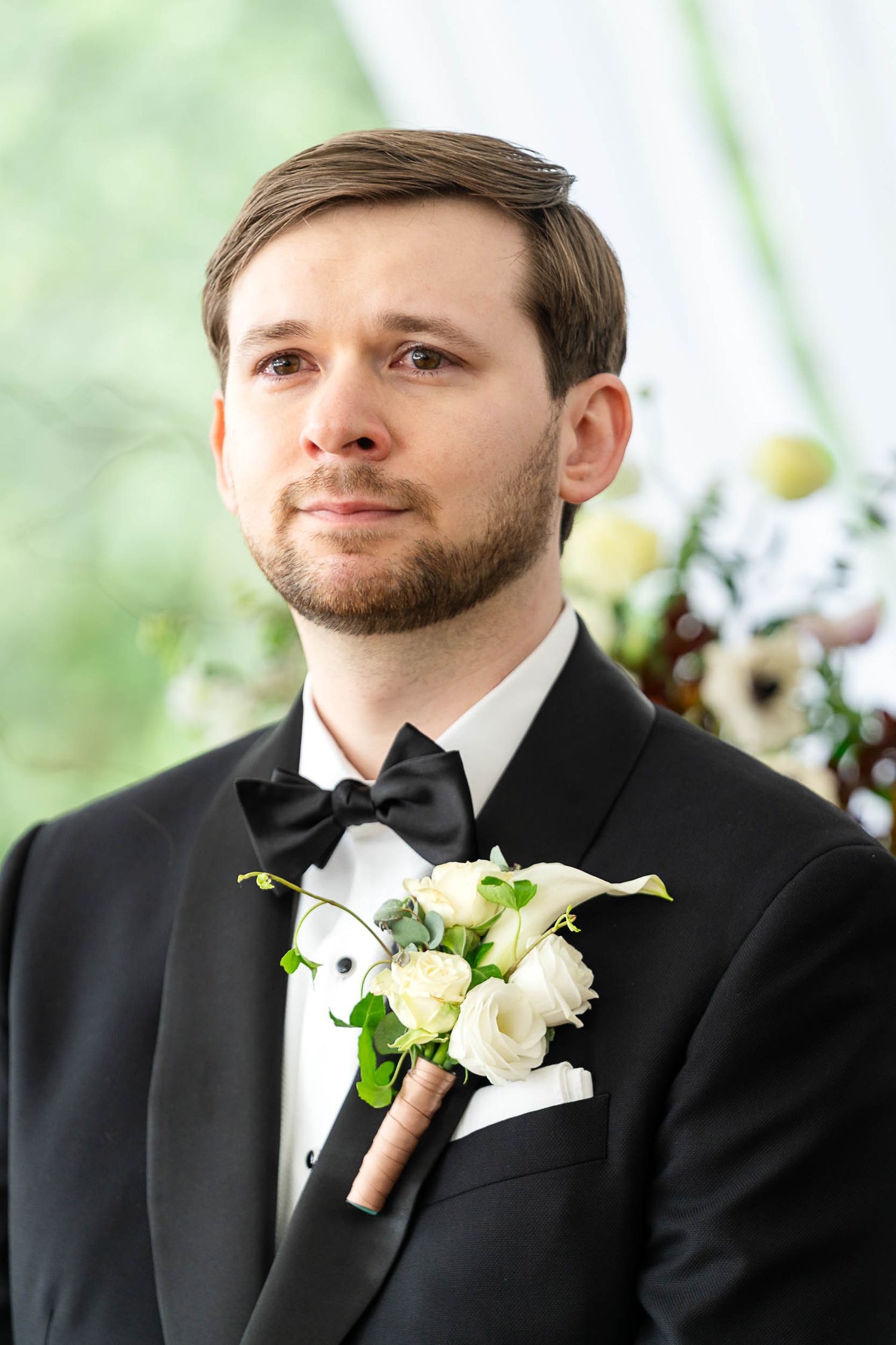 Groom in black tuxedo with white rose boutonniere and bow tie, standing at wedding ceremony altar.
