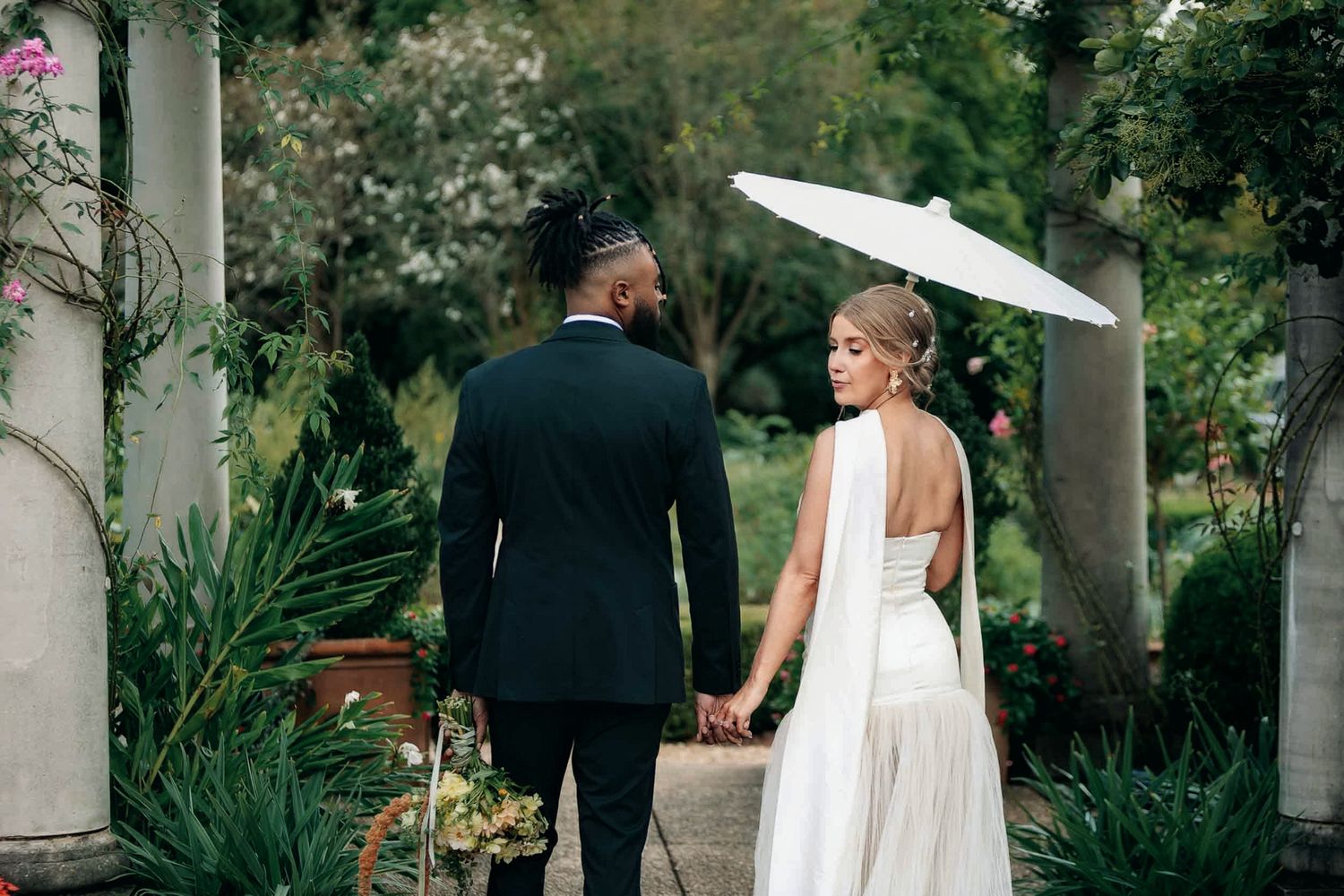 A couple in wedding attire holds hands walking through a lush garden, bride holding a white parasol.
