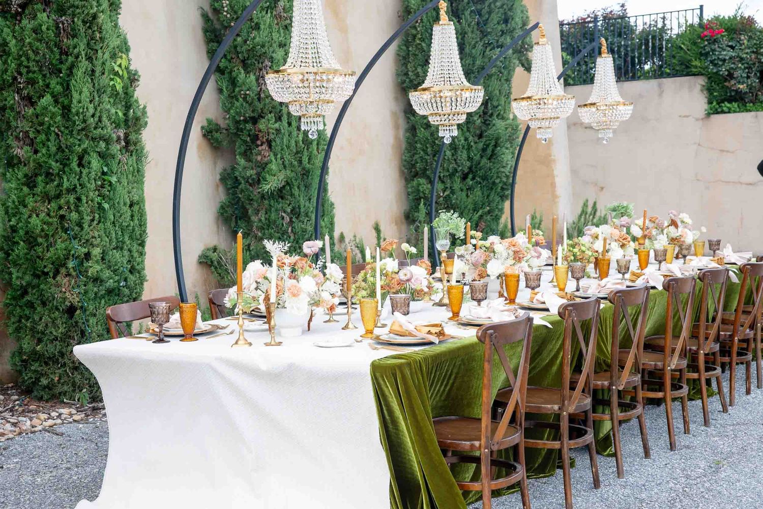 Elegant outdoor wedding reception table with green velvet runner, crystal chandeliers, gold candles, and lush greenery backdrop.