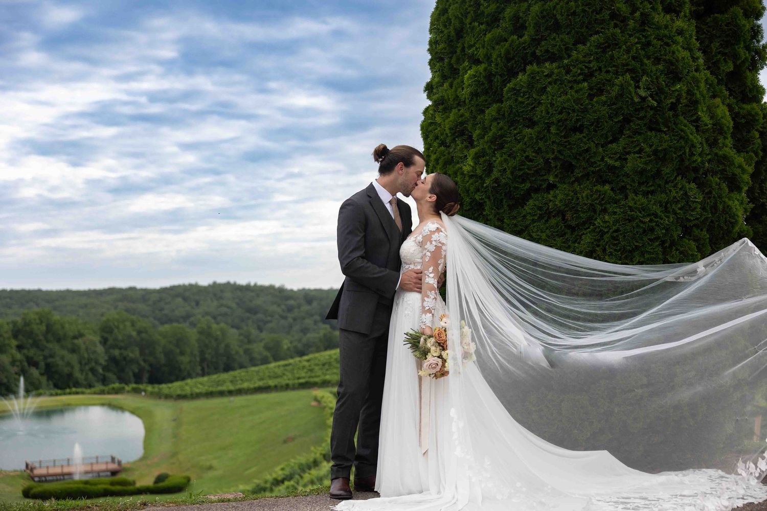 Bride and groom share a romantic kiss outdoors at a vineyard wedding venue with lush greenery and flowing veil.