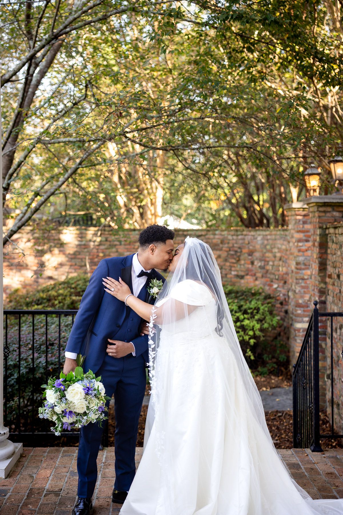 A bride and groom share a romantic kiss outdoors, surrounded by lush trees and a brick wall backdrop.