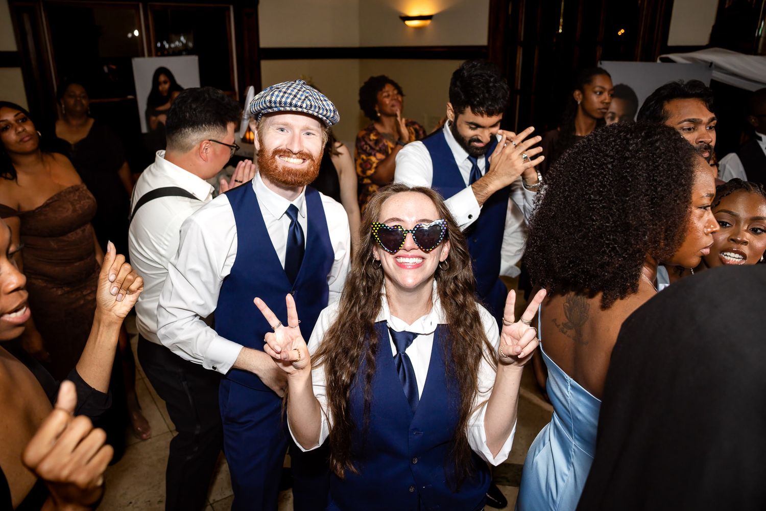 Two people in matching blue vests and hats dance and pose with peace signs at a lively indoor party.