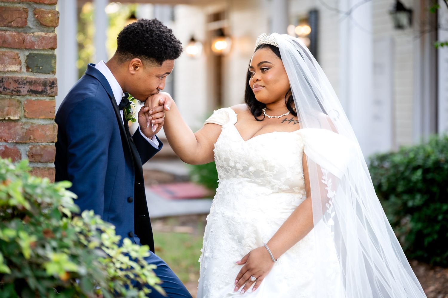 Groom in navy suit kisses bride's hand as she wears a white lace wedding gown and veil outdoors.