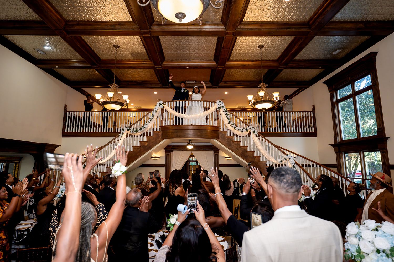 Elegant wedding reception grand staircase entrance with guests cheering as bride and groom descend in a beautifully decorated ballroom.