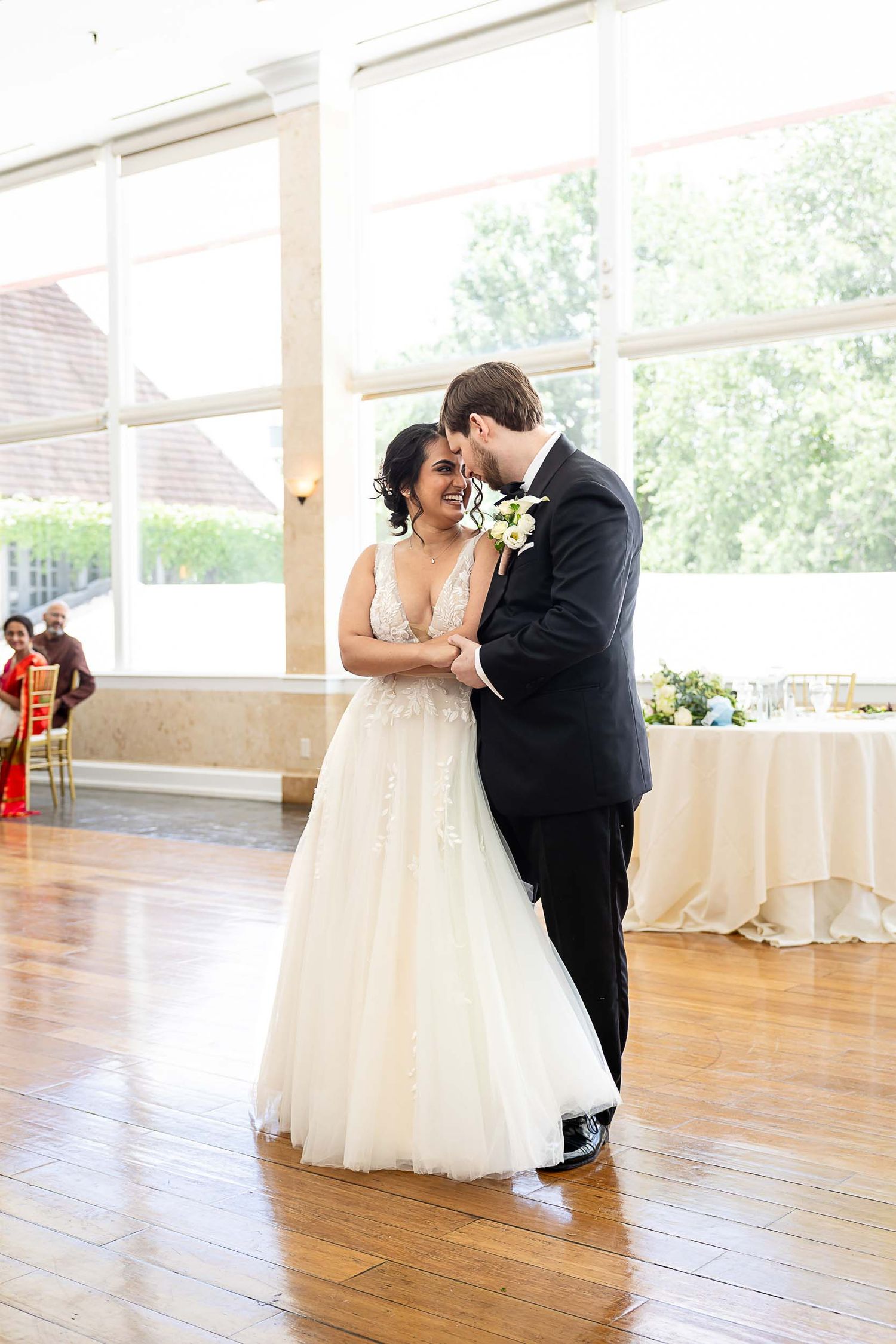 Bride and groom share their first dance in a sunlit ballroom, bride wearing a flowy white gown with floral details.