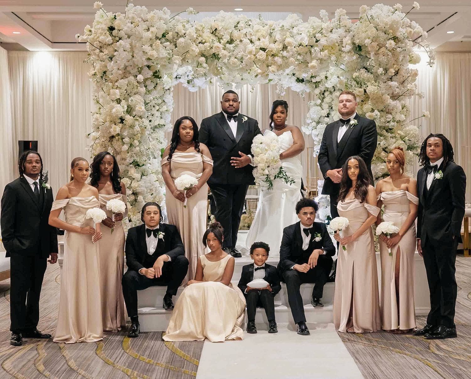 Wedding party poses under floral arch with bride and groom in center, bridesmaids in champagne gowns and groomsmen in black tuxedos.