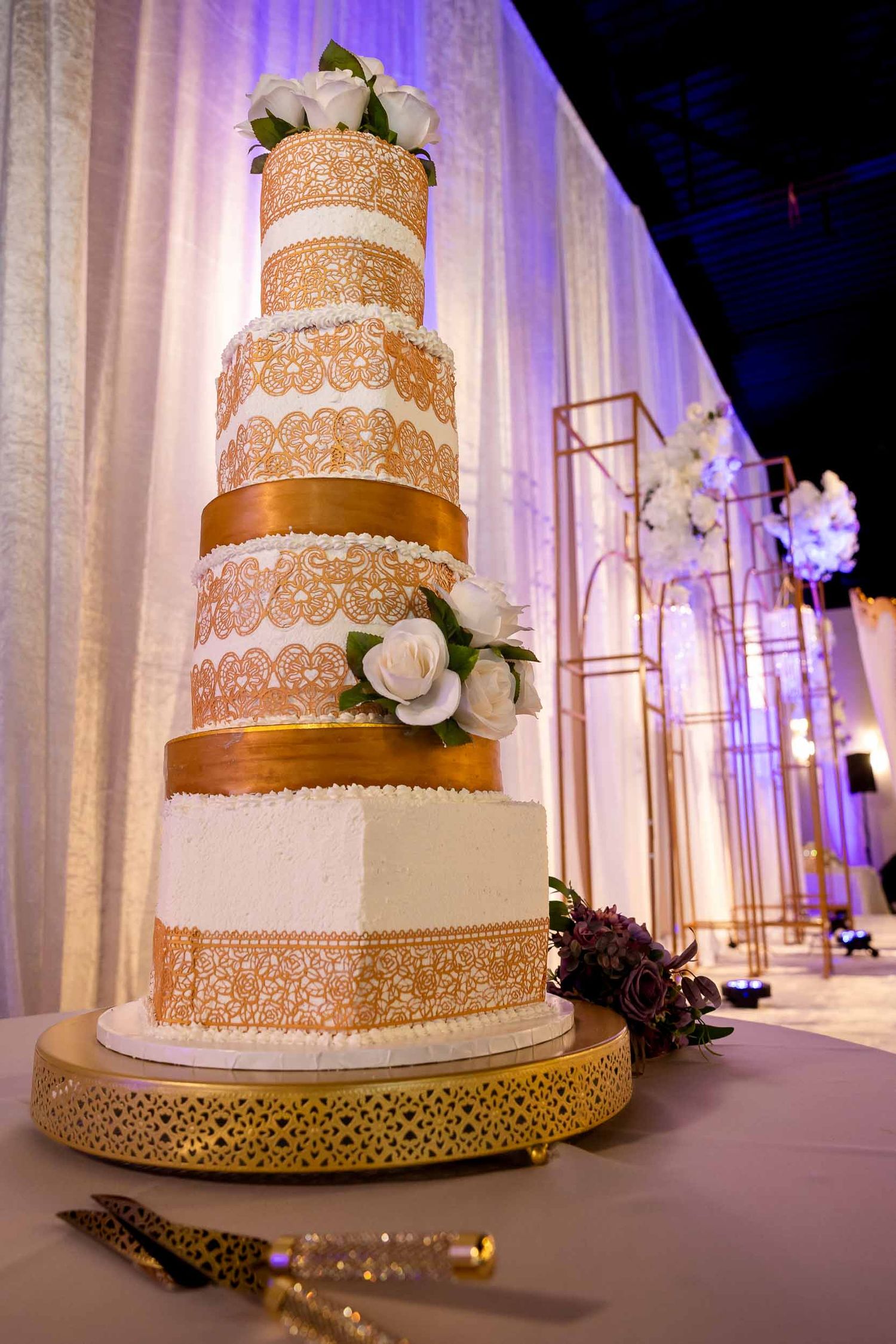 Six-tier wedding cake with gold lace detailing and white roses on a gold stand with purple ambient lighting.