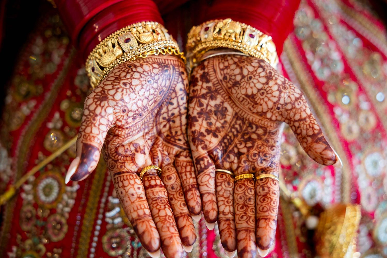 Detailed henna mehndi designs adorn palms against red traditional wedding attire background.