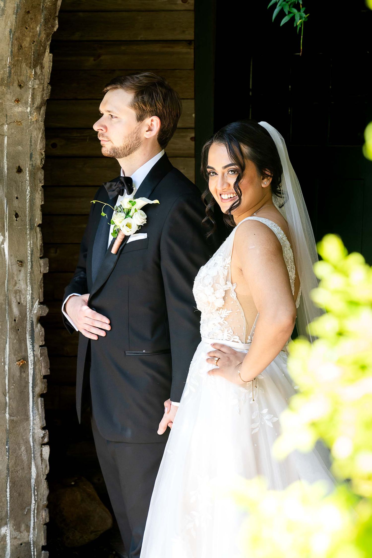 Bride in lace gown and groom in black tuxedo pose together at rustic wooden barn doorway on wedding day.